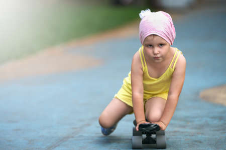 A little girl gets acquainted with a skateboard. She moves on a skateboard crouching on one knee. selective focus.の写真素材