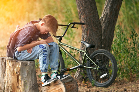 Russia, Tatarstan, August 9, 2021: a teenager is sitting on a stump in a pine forest. He talks on the phone, there is a bicycle nearby.の写真素材
