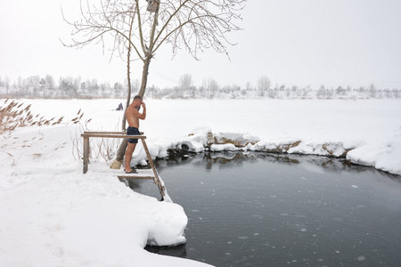 Russia, Tatarstan, January 19, 2022: frozen pond. Christening. bathing. A man in front of an ice hole is preparing to dive into icy water. Snow is falling.のeditorial素材