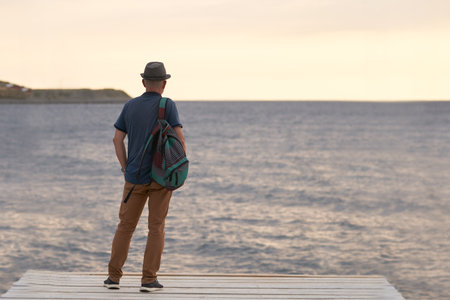 An older, gray-haired man with a hat and a backpack behind his back looks out from the pier....