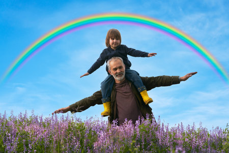 An adult man with a child on his shoulders in a spring meadow against a blue sky with a rainbow. They are playing in the airplane. copyspace.の写真素材