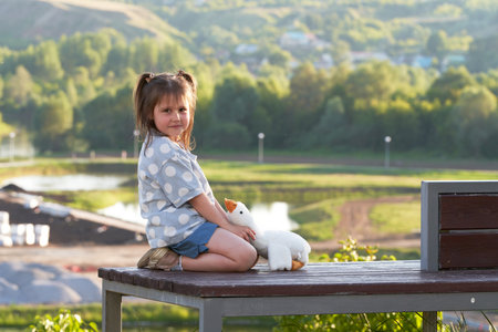 Cute little girl with a soft toy - a goose on a bench on a blurry background of nature with a pond.の写真素材