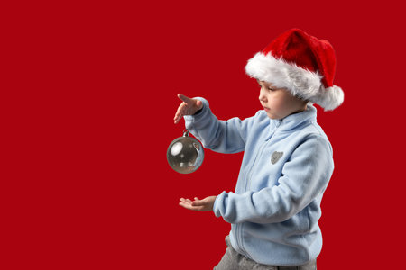 Child in Santa Claus hat holding a Christmas tree glass ball in his hand by a string. The background is red. Copy space.の写真素材