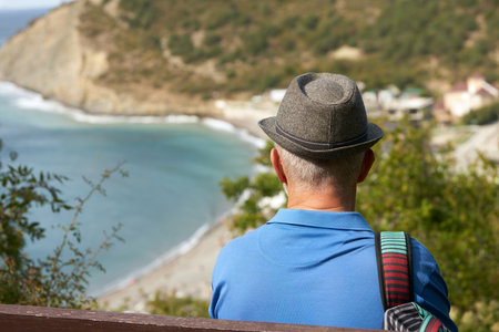 A grown man with a hiking backpack on his shoulder on a bench high up in the mountains looking down at the picturesque rocky seashore.の写真素材