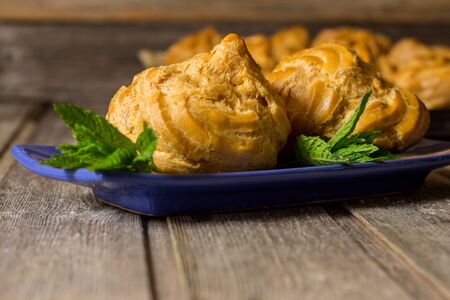 Delicious big cream puffs with cream in hand painted blue oval saucer with mint leaves on aged wooden background. Selective focus.の写真素材