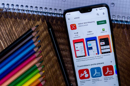 Benevento - November 29, 2020: Xmind Mind map app play store page on the display of a black mobile smartphone on wooden background. Icon log. Illustrative Editorial. In the background colored crayons, a capacitive pen and a squared notebook.のeditorial素材