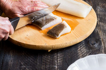 Hands of elderly woman cutting a cod fillet with knife on cutting board