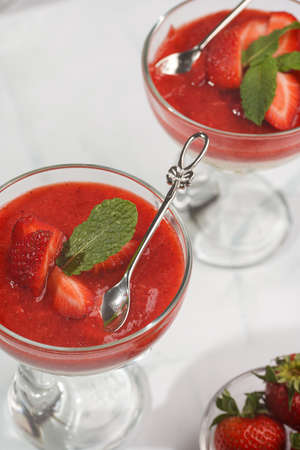 Two glasses of a traditional italian creamy pudding dessert panna cotta with peppermint and strawberry jam surrounded by fresh strawberries on a white surface, a close-upの写真素材