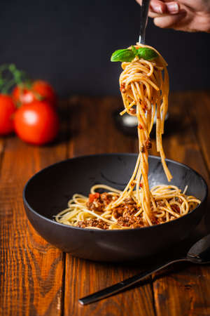 A hand holding a fork with spaghetti with bolognese tomato sauce in black bowl on wooden tableの写真素材