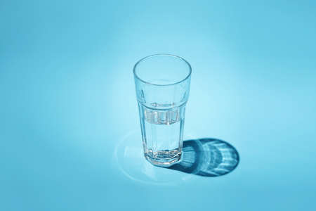 A glass of water - half full or half empty - in hard light with nice shadow on blue surfaceの写真素材