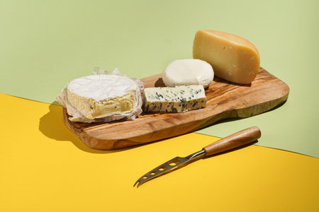 Camembert, blue cheese, mozzarella and pecorino: several cheese sorts on a wooden board on a colorful green and yellow background and a cheese knifeの写真素材