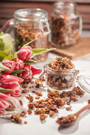 Mason jar with granola breakfast cereal with nuts. wooden spoon with scattered granola and a bouquet of tulips and two jars with granola on small wooden board in the backの写真素材