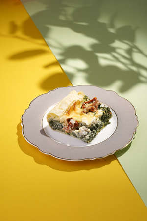 A piece of quiche, french pie filled with spinach, feta cheese and dried tomatoes on a romantic plate on a double colored (yellow and green) background in hard lightの写真素材