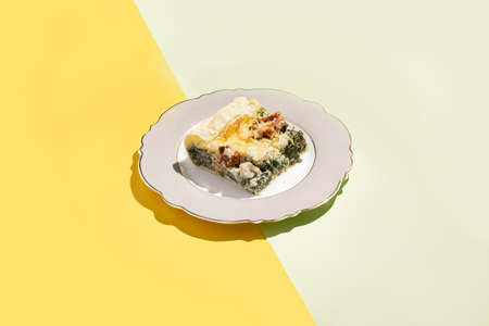A piece of quiche, french pie filled with spinach, feta cheese and dried tomatoes on a romantic plate on a seamless double colored (yellow and pastel green) background in hard lightの写真素材