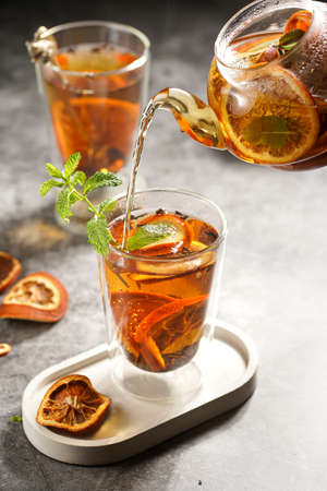 A transparent tea pot filled with black tea, oranges and mint on a gray surface pouring tea into a glassの写真素材