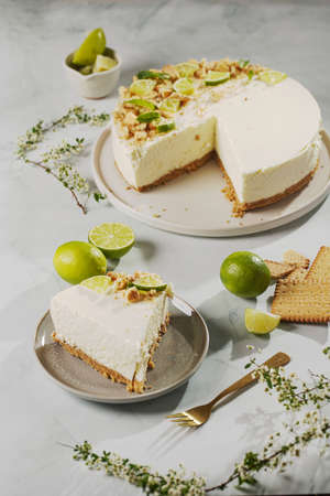 A piece of a lime cheesecake on a pastel blue surface surrounded by limes, butter cookies and flowersの写真素材