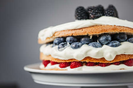 Half of layered sponge cake garnished with white cream frosting and blueberries, raspberries, blackberries, close up on gray backgroundの写真素材