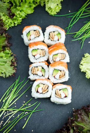 Sushi roll with salmon and avocado on black surface surrounded by greens, salad leaves and sesame seeds, flat layの写真素材