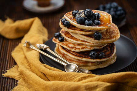 Pancakes with blueberries stacked on top of each other on black plate, two gold tea spoons and...