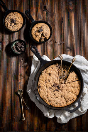 One chocolate chip cookie in a big black skillet on a kitchen towel and two small skillets with...