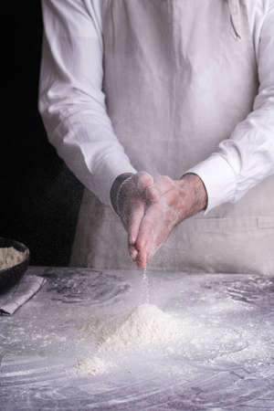 A male chef baker in a white shirt and gray a pron clapping hands with flour over a black bowl on a dark kitchen tableの写真素材
