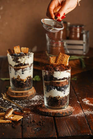 Sifting powdered sugar over two glasses with a layered dessert trifle with chocolate, cream, cookies and blueberriesの写真素材