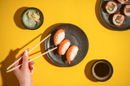 Sushi nigiri with salmon on black plate on yellow background, with soy sauce and ginger and female hand with chopsticks, top view flat-layの写真素材