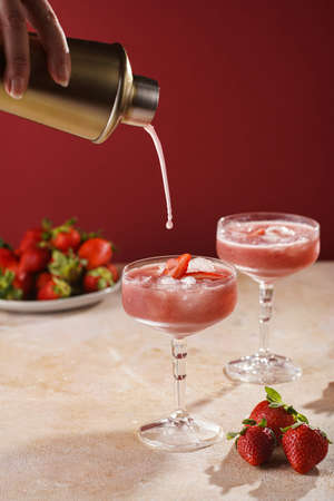 Female hand pouring cocktail our of shaker into a glass with red strawberry dessert with ice cubes and crashed ice and strawberry slices on burgundy backgroundの写真素材