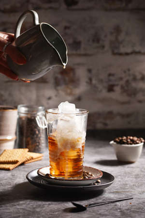 Female hand pouring milk into a glass with ice cubes and coffeeの写真素材