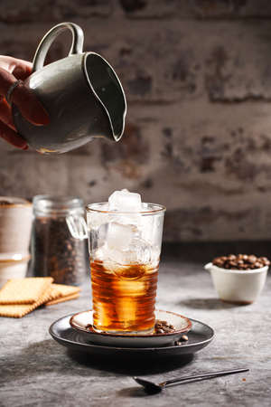 Female hand pouring milk into a glass with ice cubes and coffeeの写真素材