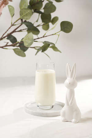 A glass of milk on a marble plate on a white table cloth, freshly baked cookies, branch of green eucalyptus, and white figurines easter bunnyの写真素材