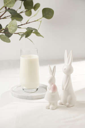 A glass of milk on a marble plate on a white table cloth, freshly baked cookies, branch of green eucalyptus, and white figurines easter bunnyの写真素材