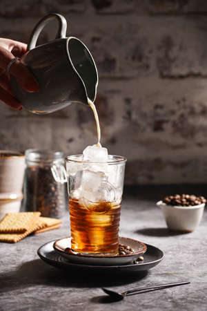 Female hand pouring milk into a glass with ice cubes and coffeeの写真素材