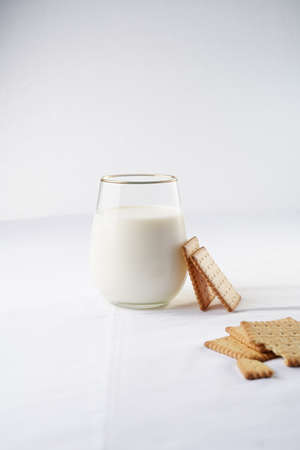 A glass of milk on the white table cloth and freshly baked cookies leaning on the glassの写真素材