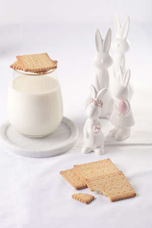 A glass of milk on a marble plate on a white table cloth, freshly baked cookies, and white easter bunnies, figurinesの写真素材