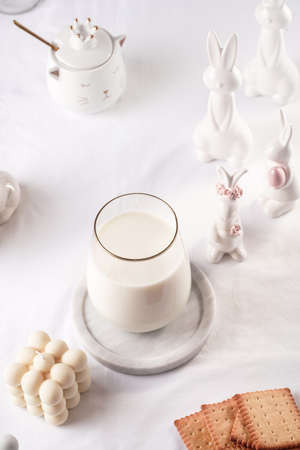 A glass of milk on a marble plate on a white table cloth, freshly baked cookies, bubble candles and white easter bunnies, figurinesの写真素材