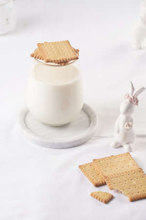 A glass of milk on a marble plate on a white table cloth, freshly baked cookies, and white easter bunnies, figurinesの写真素材