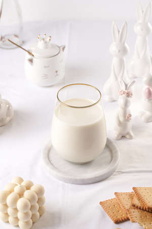 A glass of milk on a marble plate on a white table cloth, freshly baked cookies, creamy bubble candle and white easter bunnies, figurinesの写真素材