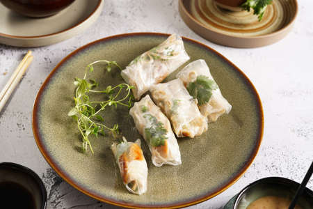 Several spring rolls with chicken, carrot and bamboo sprouts in rice paper on green plate, sesame sauce and coriander on concrete surfaceの写真素材