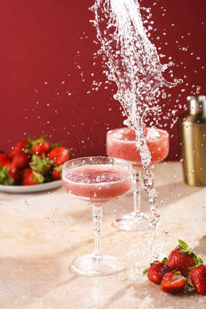 Splash of water pouring into a glass with red strawberry dessert with ice cubes and crashed ice and strawberry slices on burgundy backgroundの写真素材