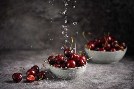 Pouring water over fresh sweet and sour dark red cherries in two gray scandi bowls on gray background and making water dropsの写真素材