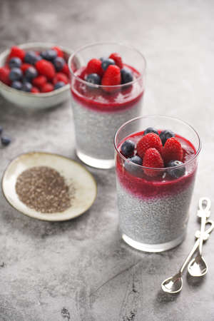 Two glasses with chia seeds pudding, raspberry jam and fresh raspberries and blueberries on a gray surfaceの写真素材