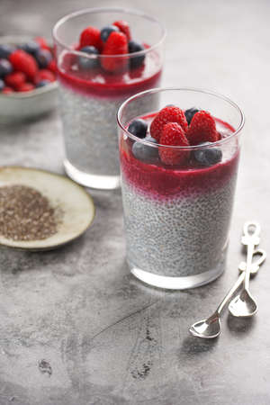 Two glasses with chia seeds pudding, raspberry jam and fresh raspberries and blueberries on a gray surfaceの写真素材