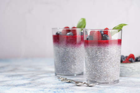Two glasses with chia seeds pudding, raspberry jam and fresh raspberries and blueberries on a concrete tray on gray surfaceの写真素材