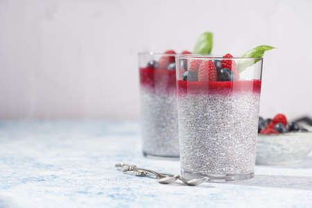 Two glasses with chia seeds pudding, raspberry jam and fresh raspberries and blueberries on a concrete tray on gray surface