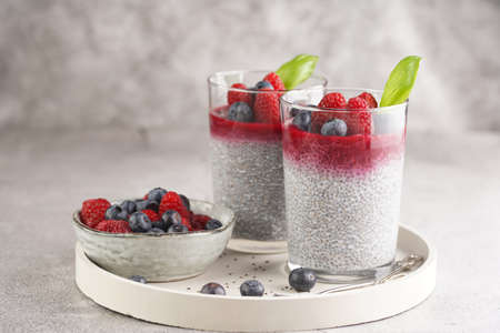Two glasses with chia seeds pudding, raspberry jam and fresh raspberries and blueberries on a concrete tray on gray surfaceの写真素材