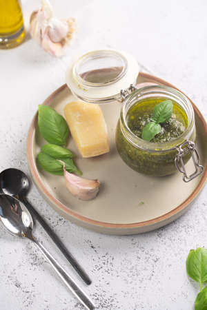 A small mason jar with green italian basil sauce pesto on a gray plate with hard cheese pecorino, garlic cloves and fresh basil leavesの写真素材