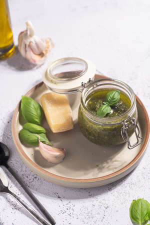 A small mason jar with green italian basil sauce pesto on a gray plate with hard cheese pecorino, garlic cloves and fresh basil leavesの写真素材