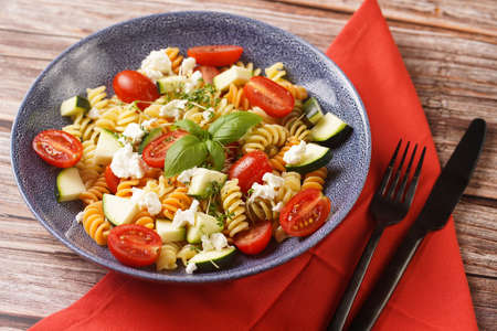 Italian pasta fusilli tricolore with cherry tomatoes, zucchini and feta cheese in a blue shallow bowl on a red napkin with black silverware on a wooden surfaceの写真素材