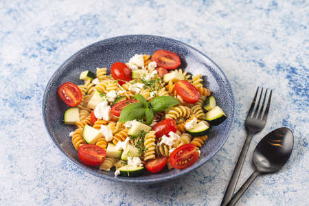 A gray cat smelling on Italian pasta fusilli tricolore with cherry tomatoes, zucchini and feta cheese in a blue shallow bowl with black silverware on a blue surfaceの写真素材